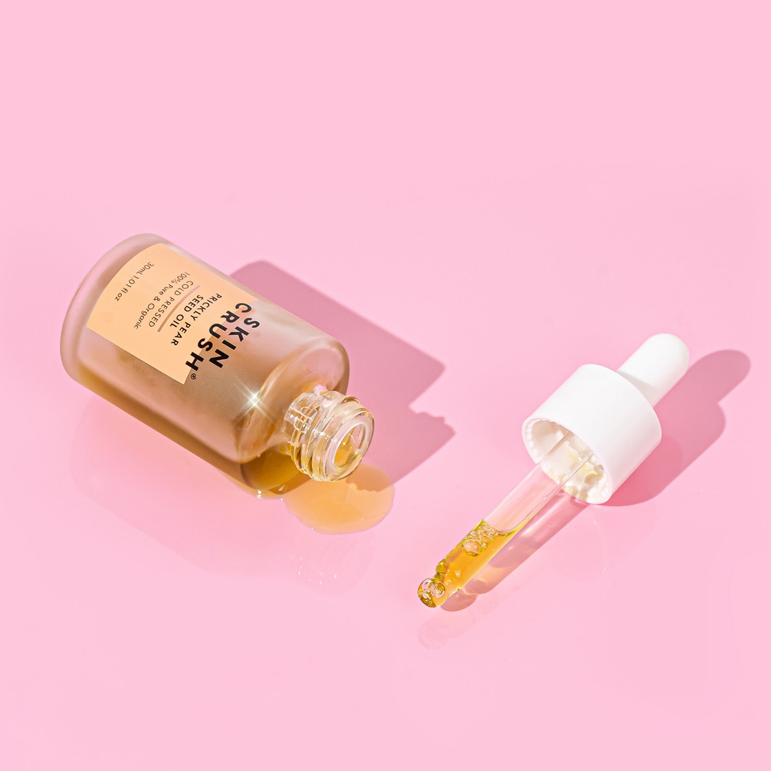 skin crush organic prickly pear seed oil spilling out of bottle on pink background with pipette and oil